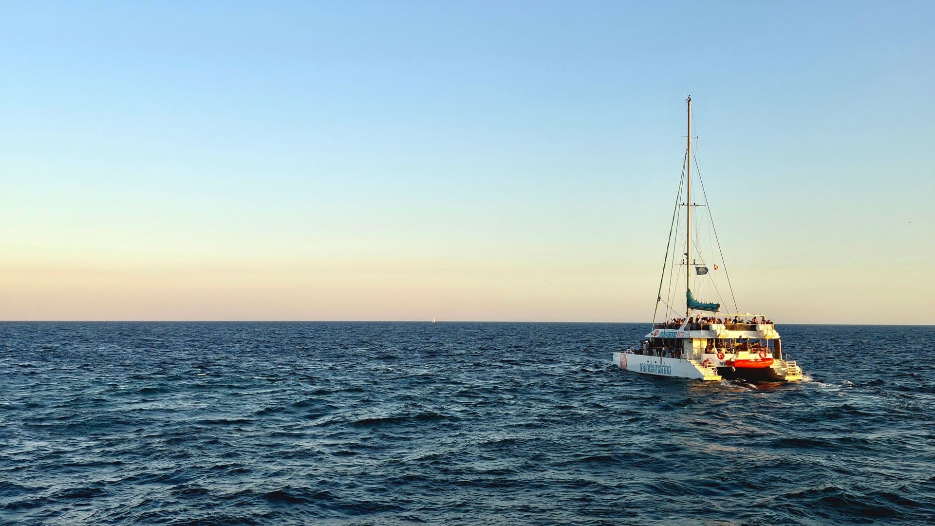 A white sailing catamaran with passengers on deck heads across the open sea under a clear sky